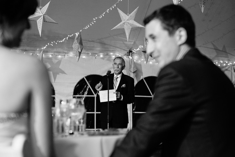 layered image, bride and groom in foreground, while father of the bride delivers toast