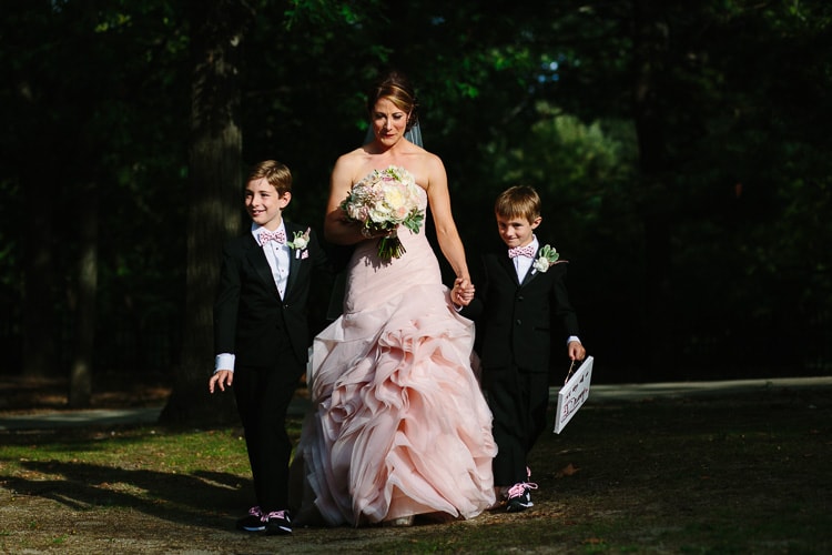 bride in pink dress walks down the aisle with her sons, wearing a pink wedding dress