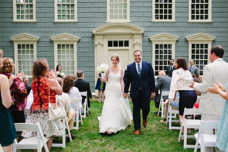 Bride and groom walk up the aisle in front of the Royall House in Medford, MA. Image by Kelly Benvenuto.