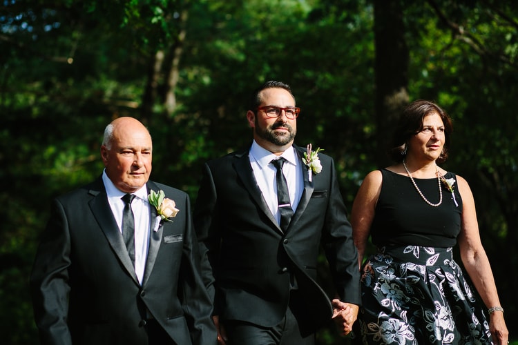 Groom walks down the aisle with his parents in dramatic light. Vibrant wedding photography in Boston by Kelly Benvenuto.
