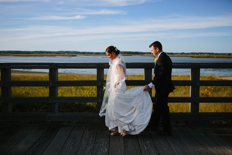 Bride and groom walk along Powder Point Bridge in Duxbury, MA while taking wedding portraits. Image by Kelly Benvenuto.