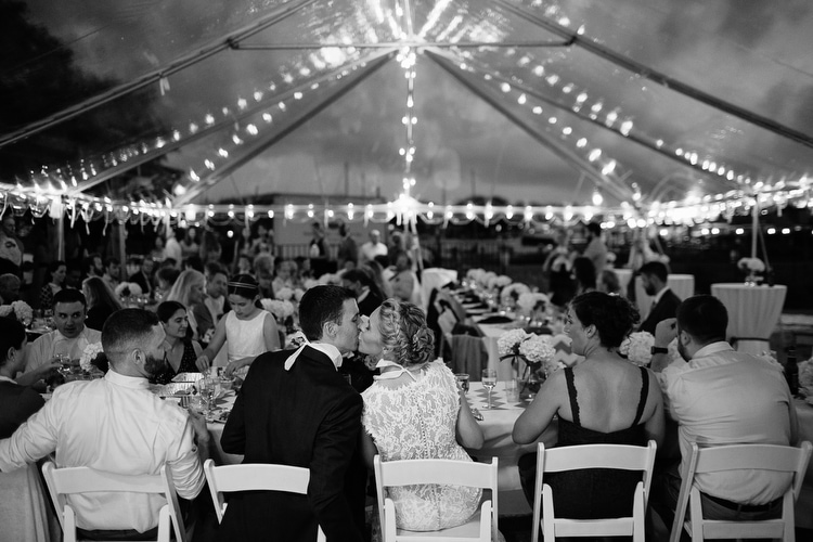 Bride and groom share a kiss under twinkle lights and clear tent at the Cape Cod Maritime Museum in Hyannis, MA. Candid wedding photography by Kelly Benvenuto.